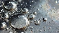 Macro photography of water droplets on dark metallic surface creating reflective spheres with light bokeh effects in background.