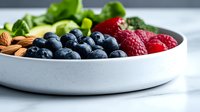 Fresh mixed berries, almonds and green salad leaves arranged in modern white ceramic bowl on light background, healthy breakfast concept.