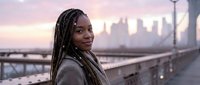 Young African American woman with braids smiling at sunset on urban bridge with city skyline background. Professional portrait for lifestyle and diversity content.