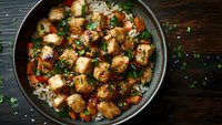 Spicy chicken pieces with vegetables and herbs served over steamed rice in dark bowl, garnished with fresh parsley on rustic wooden background.