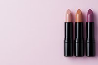 Three matte lipsticks in nude pink, caramel and plum shades with black tubes arranged on soft pink background. Minimal beauty flat lay composition.
