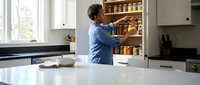 African American woman organizing spice jars in modern kitchen pantry cabinet with white countertops and natural lighting from window.