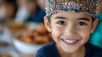 Young Asian boy wearing traditional songkok cap with blue and gold pattern smiles brightly at camera during Eid celebration with family gathering and festive food in background.