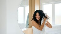 Young Hispanic woman with curly hair drying hair with towel in modern bathroom mirror reflection morning routine self-care wellness lifestyle.