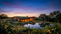 Modern luxury home with warm lighting reflected in tranquil pond at sunset, surrounded by native landscape and mature trees in serene natural setting.