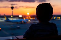 Silhouette of young boy watching sunset at airport terminal with control tower and airplanes on tarmac, orange sky and city lights creating dreamy atmosphere.