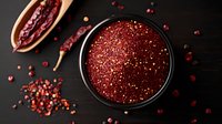 Ground pink peppercorns in black ceramic bowl on dark wooden background with scattered whole peppercorns and dried pods, dramatic food styling.