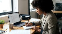 Young African American businesswoman working at modern office desk with laptop, taking notes and reviewing documents in professional workspace.