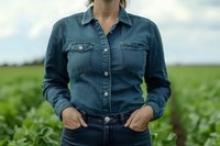 Young woman in denim shirt and jeans standing in agricultural field with green crops, hands in pockets, casual countryside fashion style for farm lifestyle.