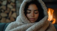 Young African American woman wrapped in cozy sherpa blanket relaxing with closed eyes by fireplace, warm evening atmosphere creates peaceful winter mood.