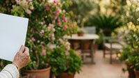Senior adult hand holding blank white card in lush garden with flowering plants and terracotta pots, suitable for greeting cards, invitations, and gardening promotions.