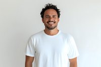 Hispanic man with curly dark hair and beard wearing white t-shirt smiling confidently against light background. Natural genuine expression shows joy and optimism.