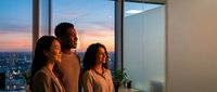 Young diverse group of three people standing together at floor-to-ceiling windows in modern high-rise office, gazing at sunset cityscape view. Corporate team enjoying evening moment.