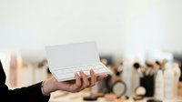 Businessperson holding open white business card box in professional office environment with blurred colleagues in background for corporate branding and networking purposes.