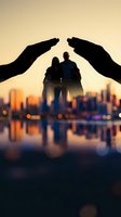Protective hands sheltering silhouetted couple against golden sunset city skyline. Insurance, safety, security and care concept for family protection services.