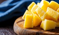 Fresh yellow mango chunks cut into cubes arranged on rustic wooden cutting board against dark blue textile background, close up view with selective focus.
