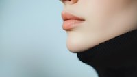 Close-up profile view of feminine lips and chin against soft blue background, wearing black turtleneck. Minimalist beauty and fashion composition with gentle lighting.
