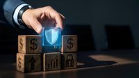Businessman hand protecting glowing shield icon above wooden blocks with financial symbols representing security and risk management in corporate finance.
