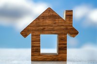 Wooden house silhouette against blue cloudy sky background. Simple minimalist home shape symbol represents real estate, property ownership and residential concepts.