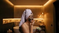 Happy young African American woman applying face mask in luxury spa bathroom with warm ambient lighting for skincare routine and wellness relaxation.