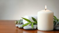 Lit white candle with green bamboo leaves and smooth stones on wooden surface creating peaceful spa atmosphere for relaxation and wellness.
