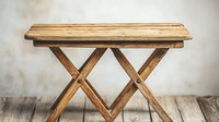 Rustic wooden folding table with crossed legs on weathered wooden floor against textured wall background, vintage farmhouse style furniture piece.