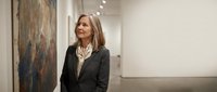 Professional middle-aged Caucasian businesswoman in dark suit standing in modern art gallery with abstract paintings and white walls.