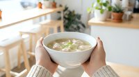Asian woman holding warm miso soup bowl with tofu and green onions in bright kitchen. Comfort food for wellness and healthy eating lifestyle.