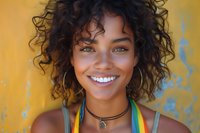 Young African American woman with curly hair smiling brightly wearing colorful summer top against vibrant yellow wall background for lifestyle content.