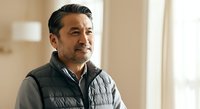 Middle-aged man with dark hair and facial hair wearing a black quilted vest over a gray long-sleeve shirt, standing indoors with blurred background, suitable for business, lifestyle, and professional content.