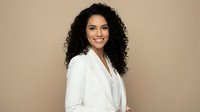 Professional Hispanic businesswoman with curly hair in white blazer smiling confidently against neutral beige background for corporate marketing materials.