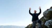 Middle-aged man with bald head and light skin tone, wearing dark athletic jacket, celebrating with arms raised on rocky mountain summit overlooking scenic valley landscape during daytime.