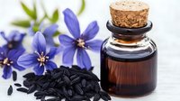 Small brown glass bottle with cork stopper containing essential oil next to fresh blue borage flowers and black seeds on white wooden background for aromatherapy.
