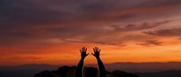 Silhouetted hands raised toward vibrant orange and purple sunset sky with mountains in background, expressing freedom, celebration, and gratitude during golden hour.