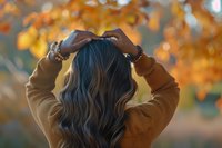 Young woman with long dark wavy hair wearing brown sweater viewed from behind against blurred autumn foliage background with warm orange bokeh lights creating cozy fall mood.