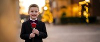 Young Caucasian boy in formal black suit holding red rose with warm bokeh lights background for romantic occasions and special events.