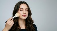 Young brunette woman applying makeup with powder brush, eyes closed, professional beauty routine on gray background for cosmetics and skincare concepts.