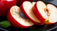 Fresh red apple slices sprinkled with black chia seeds on dark plate, green leaf garnish, macro food photography showing texture and details.