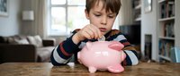 Young boy with brown hair and light complexion inserting coin into pink piggy bank on wooden table, learning financial literacy and savings habits at home.