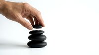 Close-up of male hand stacking smooth black stones in zen balance against white background, representing wellness, mindfulness and spa therapy concepts.