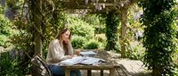 Young woman working remotely on laptop in peaceful garden pergola surrounded by lush greenery, creating productive outdoor workspace for freelancing and study.