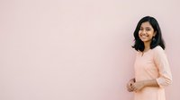 Young woman with dark hair and South Asian ethnicity wearing a light peach blouse, smiling at camera against pink background. Suitable for business, lifestyle, and portrait projects.