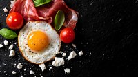 Sunny side up egg with prosciutto slices, cherry tomatoes, fresh basil leaves and crumbled feta cheese on dark stone background. Food photography for menu design.