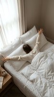 Woman stretching in bed after waking up, arms raised above head, surrounded by white pillows and bedding near bright window with sheer curtains.