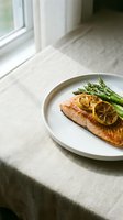 Grilled salmon fillet topped with lemon slices served with fresh asparagus on white plate, placed on linen tablecloth near window for fine dining and healthy meal concepts.