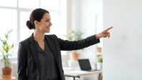 Professional businesswoman in dark suit pointing forward with confident smile in modern bright office workspace for corporate presentations.