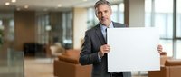 Middle-aged Caucasian businessman in dark suit holding blank white sign in modern office space for corporate messaging and professional presentations.