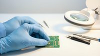 Laboratory technician in blue gloves examining green circuit board under magnifying lamp for electronic component analysis and quality control testing.