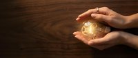 Hands protecting glowing light bulb with innovation icons on wooden background representing creative ideas and business solutions.