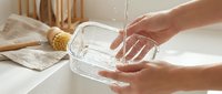 Hands washing a clear glass food container under running water in a clean kitchen sink, with a natural brush and linen towel nearby.
